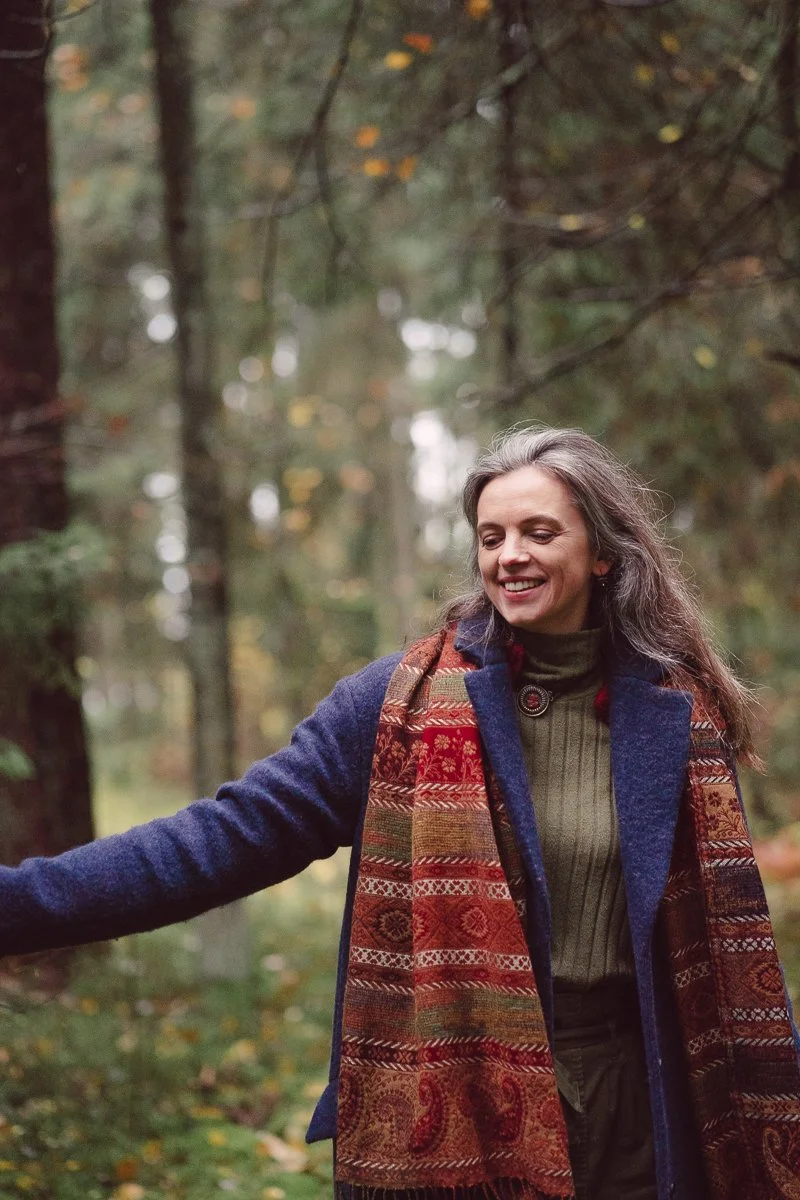 A woman with long, gray hair smiling in a wooded forest, wearing a navy blue coat, a green sweater, and a multicolored patterned scarf.