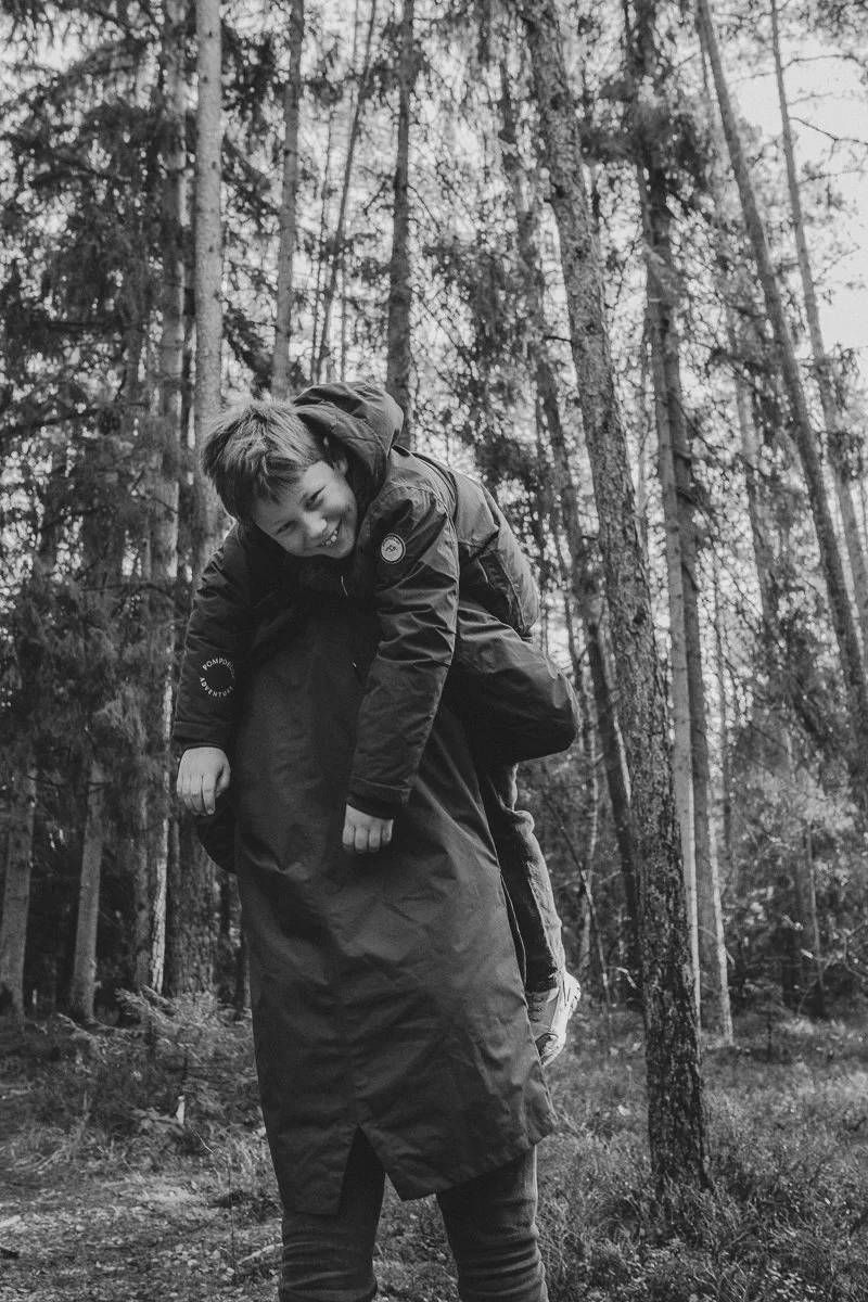 A person carrying a boy on their shoulders in a forest with tall trees, black and white photo.