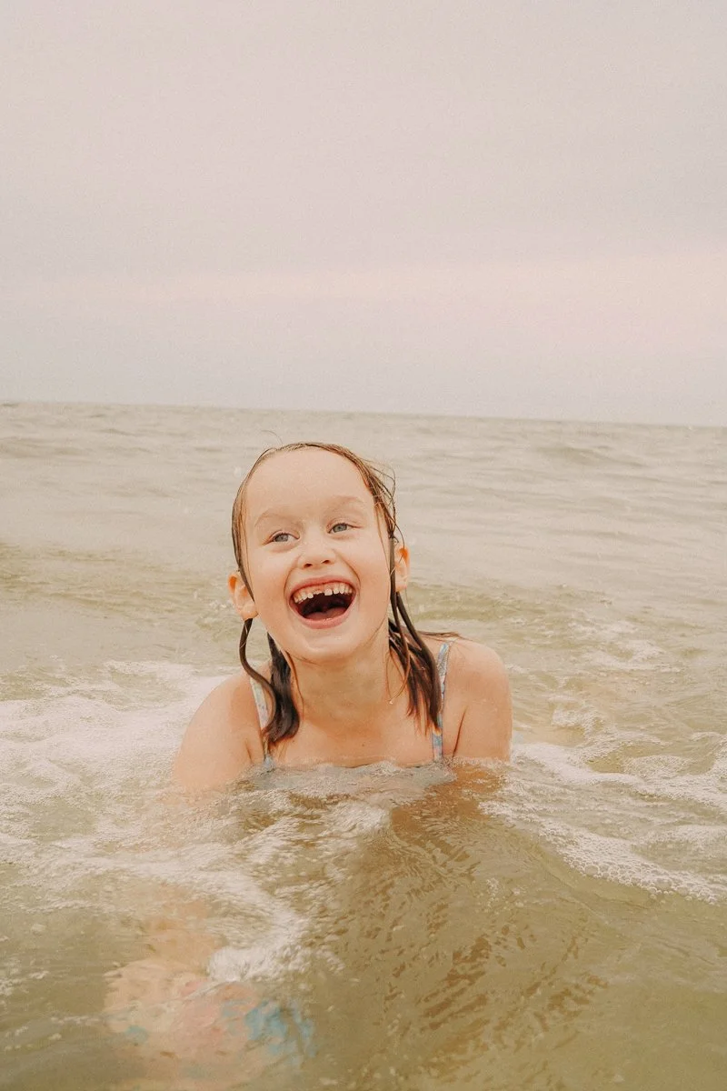 A smiling young girl with wet hair is in shallow ocean water, enjoying a day at the beach.