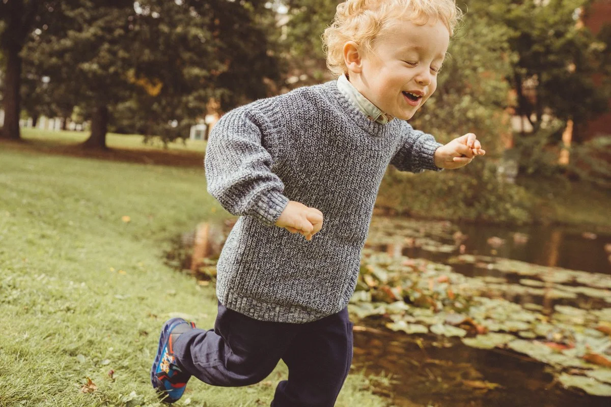 A young boy with curly blonde hair running by a pond with lily pads in a park during autumn.