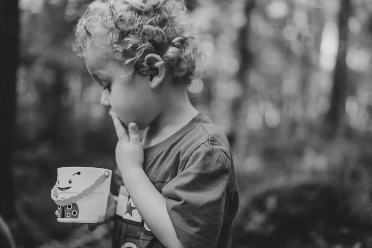 A young boy with curly hair looking at a cup with a smiley face on it, standing outdoors in a forested area.