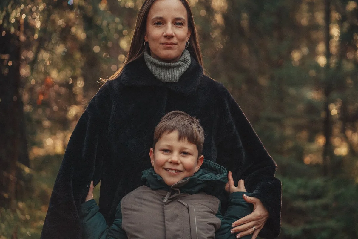 A woman and a young boy outdoors in a forest during sunset, smiling and facing the camera.