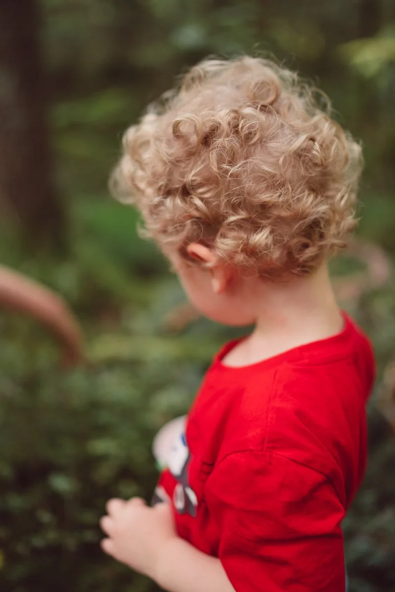 Child with curly blonde hair wearing a red shirt in a wooded outdoor setting.