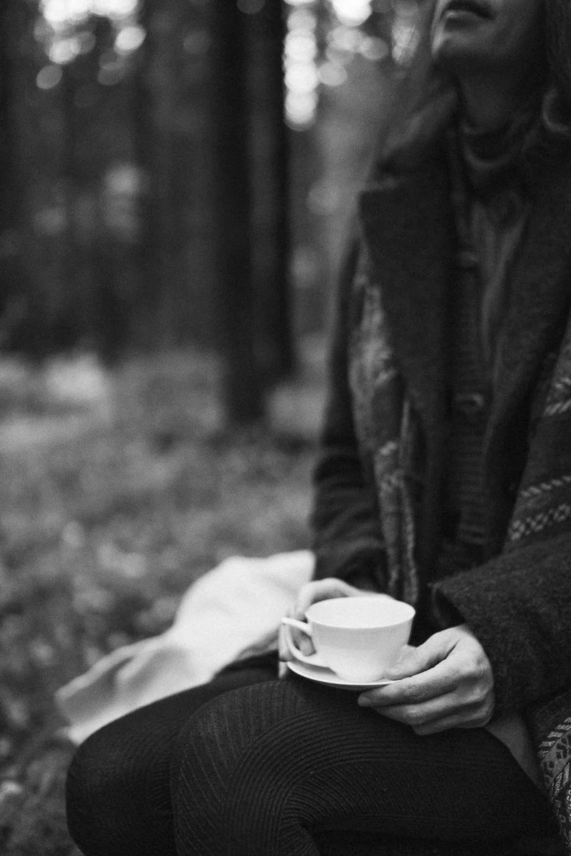 A person sitting outdoors holding a white teacup and saucer in their hand, with a blurred natural background.