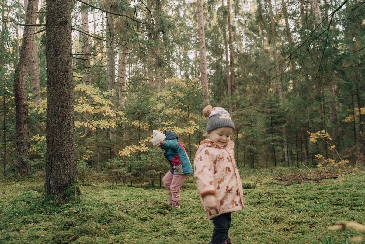 Two young children explore a forest, one smiling and the other looking downward, surrounded by trees and lush greenery.