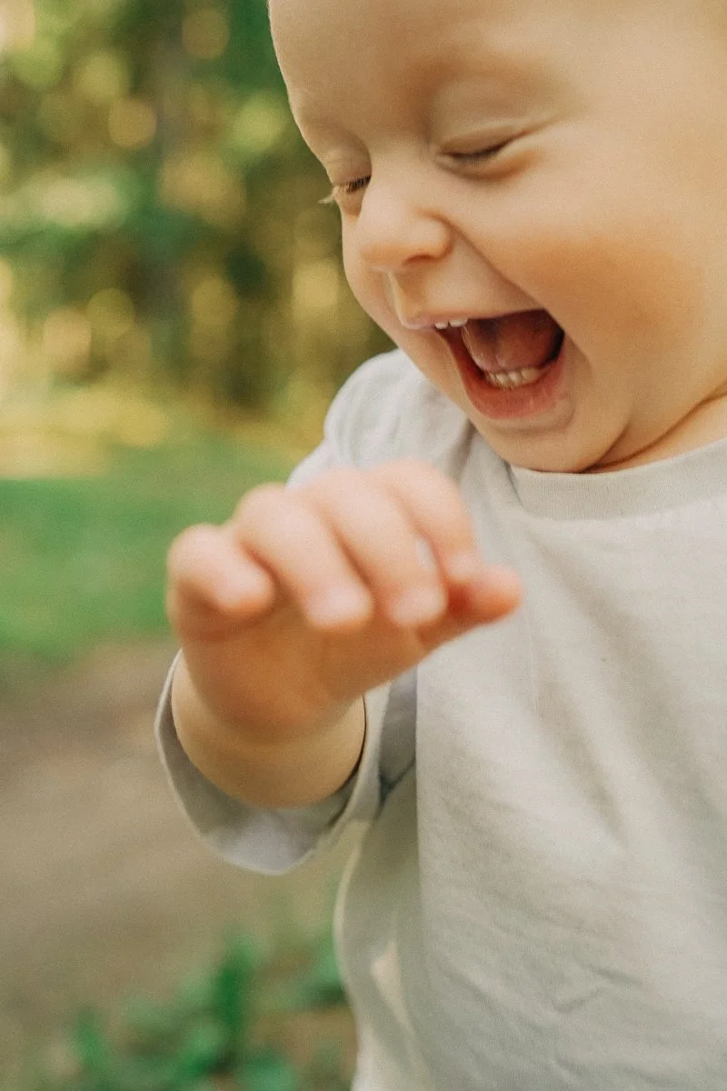 Close-up of a laughing young child with blond hair, wearing a light gray shirt, outdoors with a blurred green background.