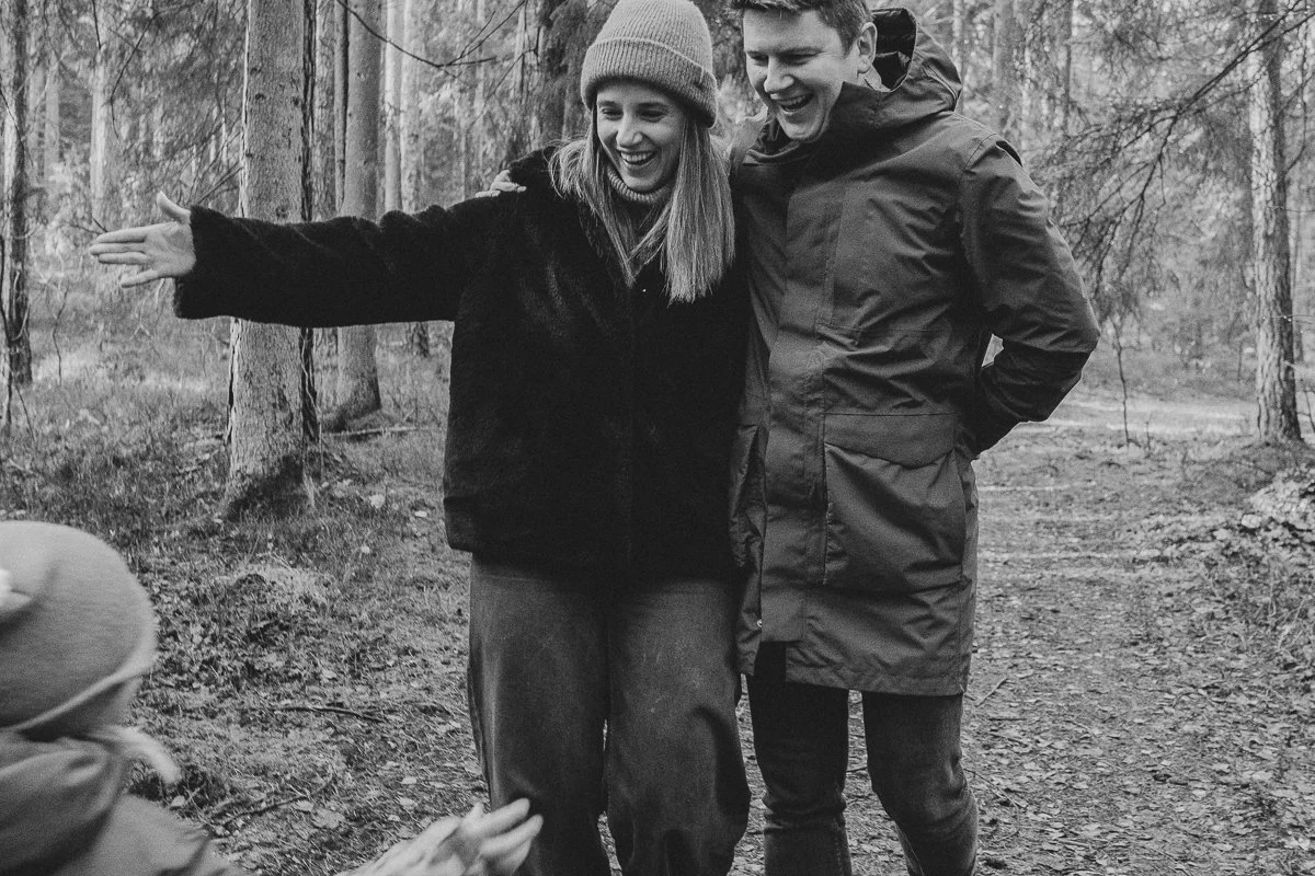 A woman and a man standing together in a wooded area, smiling and looking at each other, with the woman extending her arm.