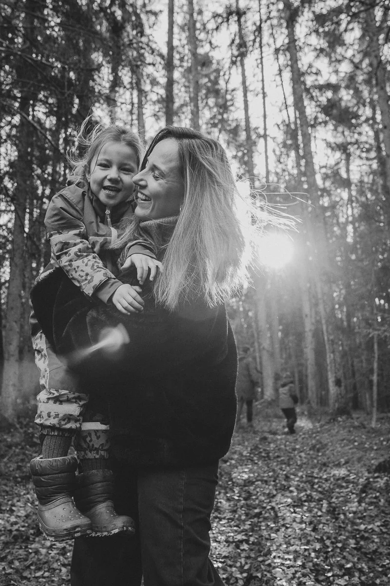 A woman holding a young girl in a forest with sunlight filtering through trees, both smiling and enjoying nature.