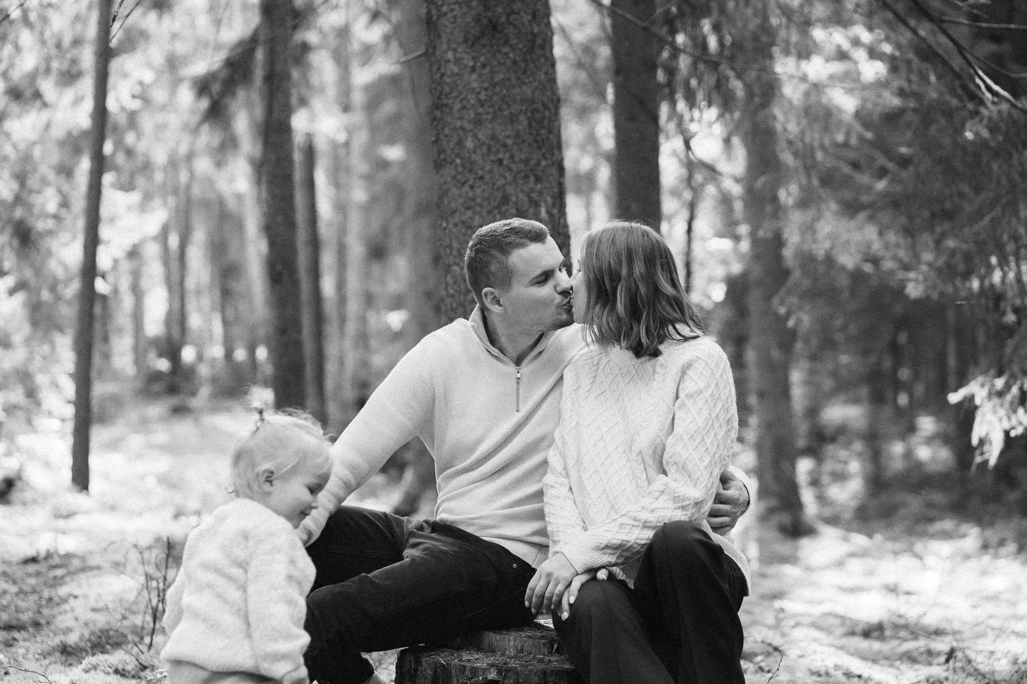 A couple sharing a kiss in a forest, accompanied by two dogs, one near their feet and the other sitting close by.