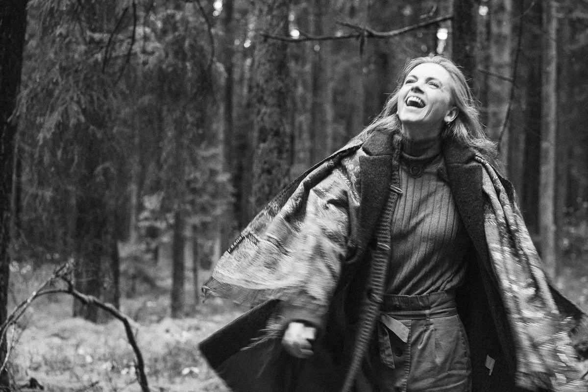 A woman smiling and laughing while walking in a wooded forest area with trees in the background, wearing a coat and plaid blanket wrapped around her shoulders.