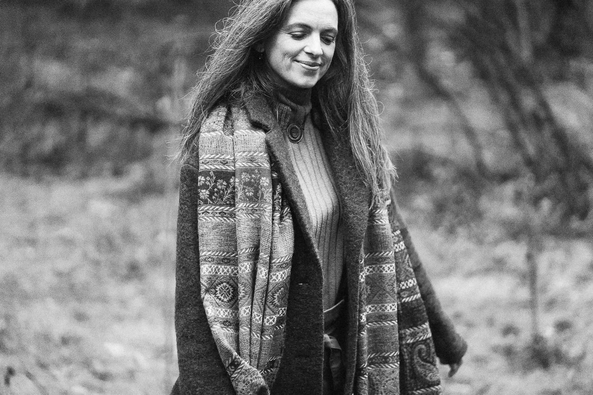 A woman with long hair smiling outdoors, wearing a patterned coat over a turtleneck sweater.