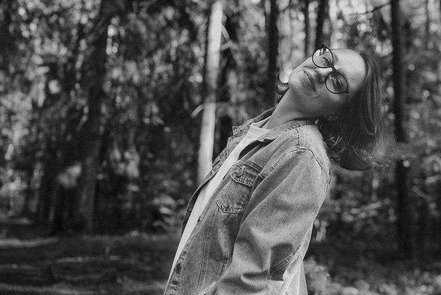 A woman with glasses and shoulder-length hair standing outdoors in a forest, smiling and leaning her head to the side.