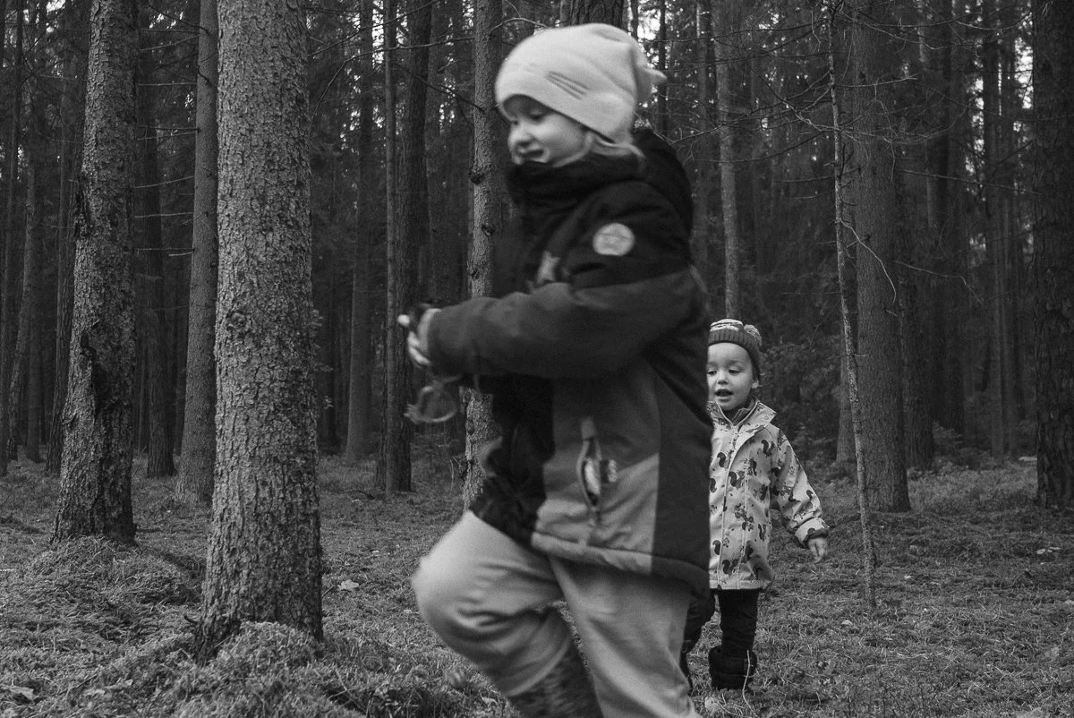 Two children hiking in a forest, one boy in the foreground wearing a winter jacket and hat, and a girl in the background also in a winter coat and hat.