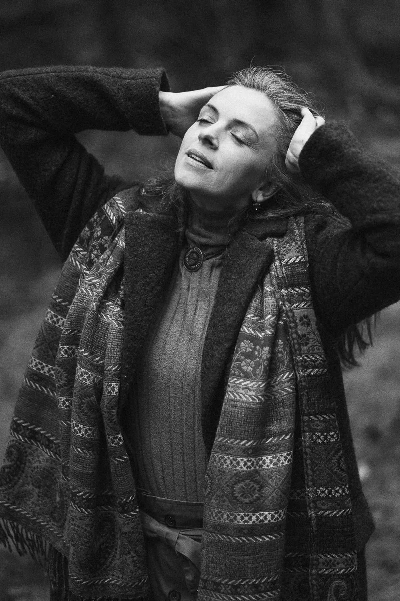 A woman with closed eyes and a relaxed expression holds her head with both hands, standing outdoors.