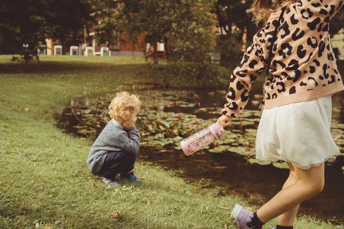 A young girl with curly blonde hair, squatting on the grass by a pond, looking at a girl with a water bottle. The girl with the water bottle is standing near the water, wearing a leopard print sweater and a white skirt, holding the bottle towards the