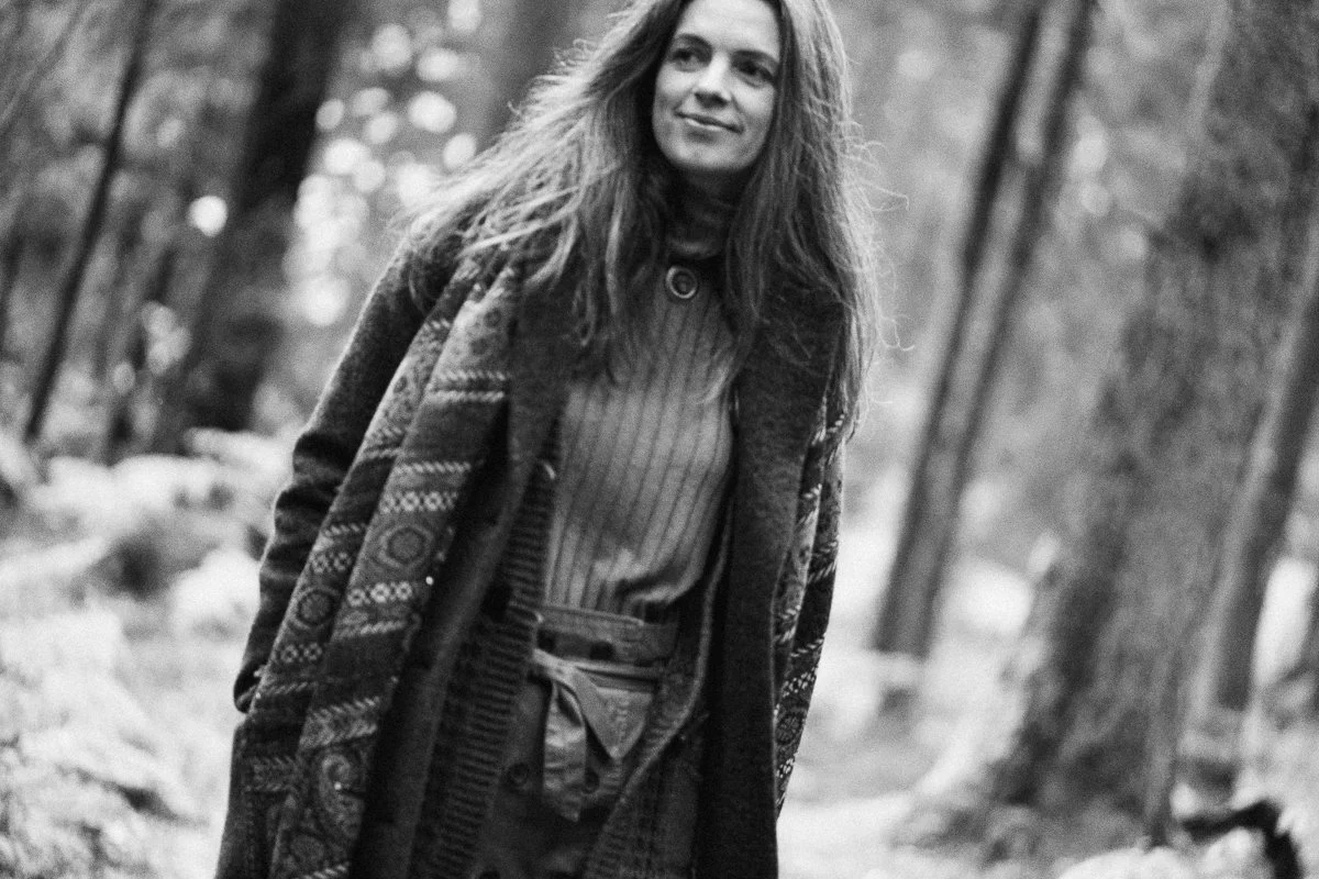 A woman with long hair outdoors in a wooded area, wearing a patterned coat and striped shirt, smiling slightly.