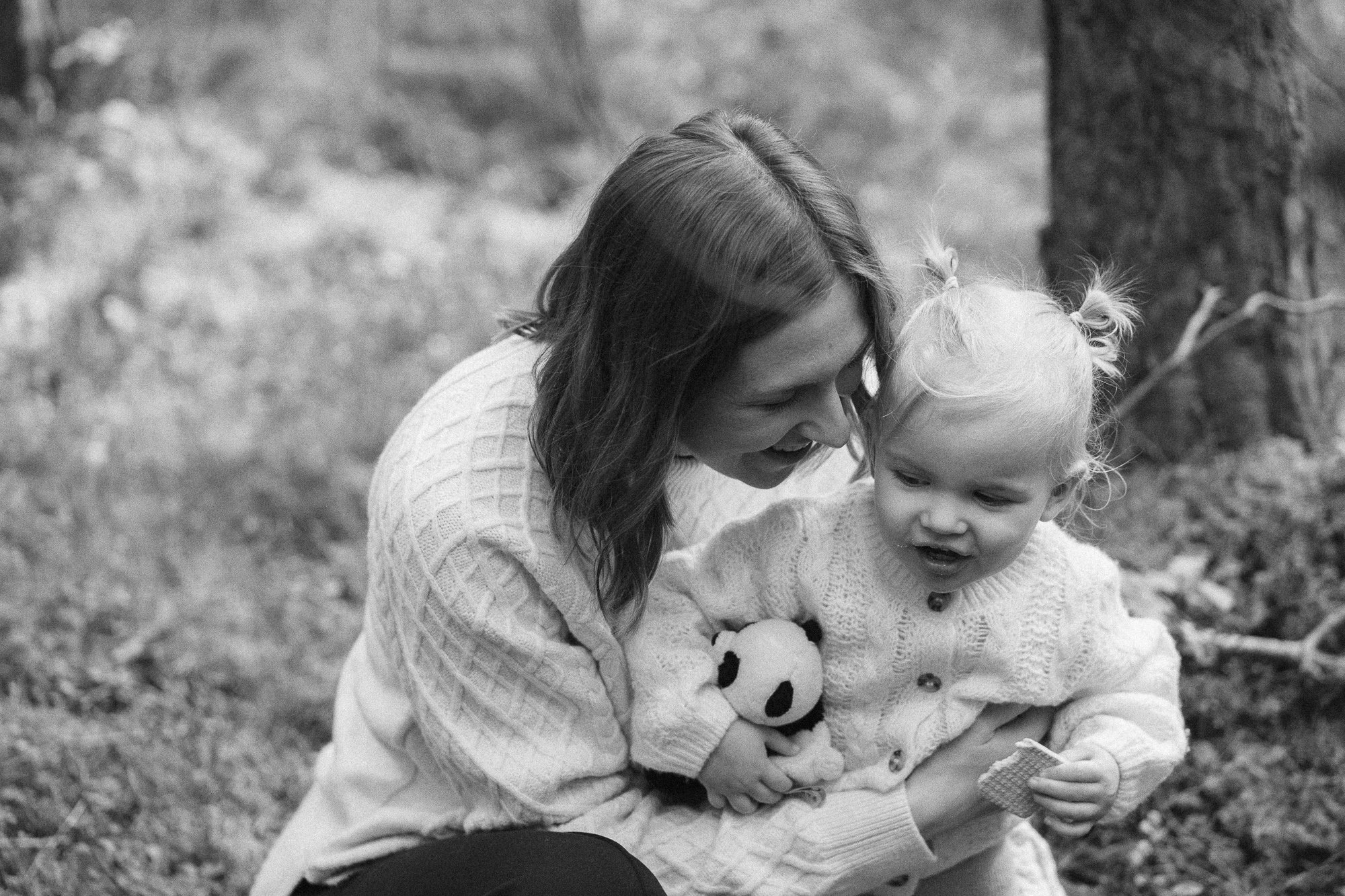 A woman and a young girl sitting outdoors on the grass, both smiling and looking at a small object the girl is holding in her hands. The woman has long hair and the girl has pigtails, both wearing sweaters.