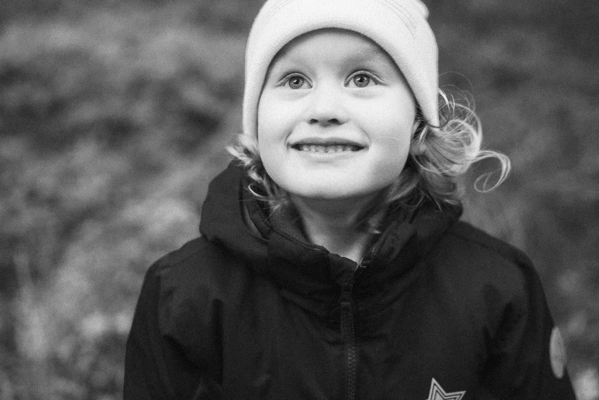A young child with curly hair and light eyes wearing a beanie and a winter jacket, looking up with a smile.