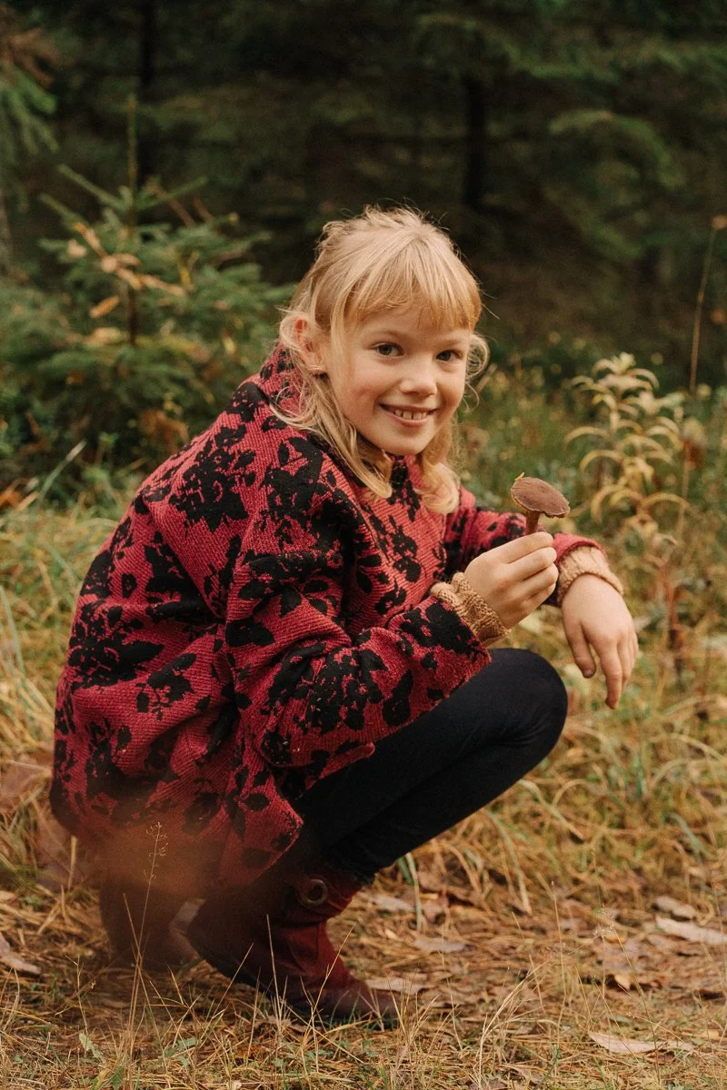 A young girl with blonde hair in a forest, crouching on the ground, holding a mushroom, smiling at the camera, wearing a red and black patterned jacket, black pants, and red boots.