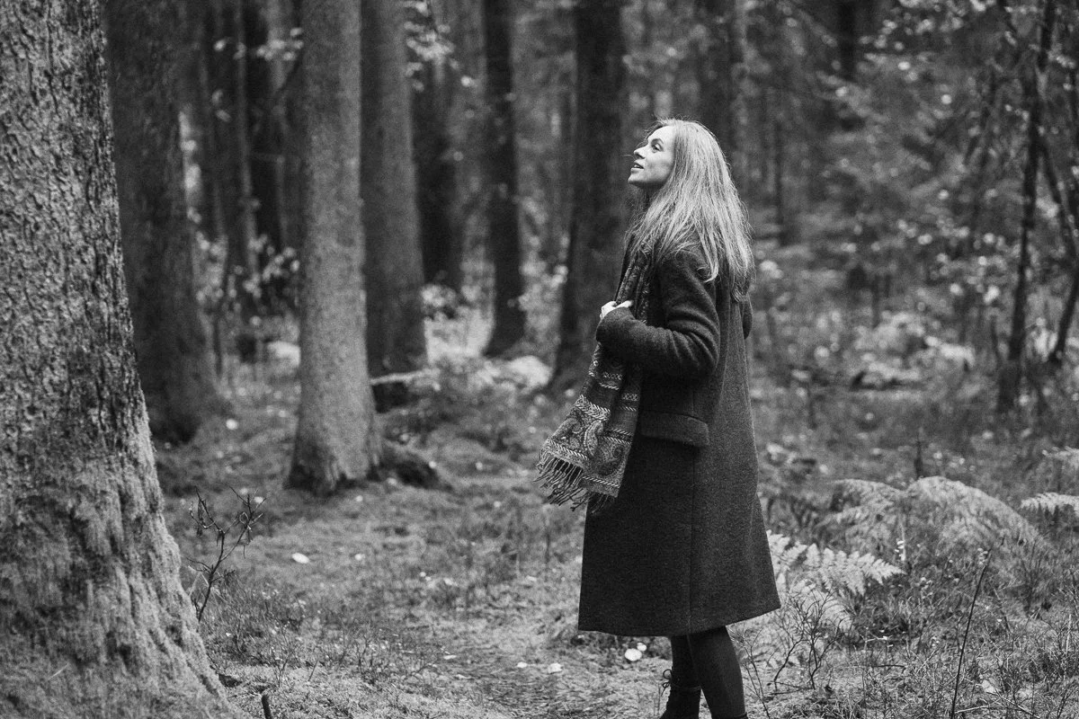 A woman standing in a forest with tall trees, wearing a long coat and scarf, gazing upward.