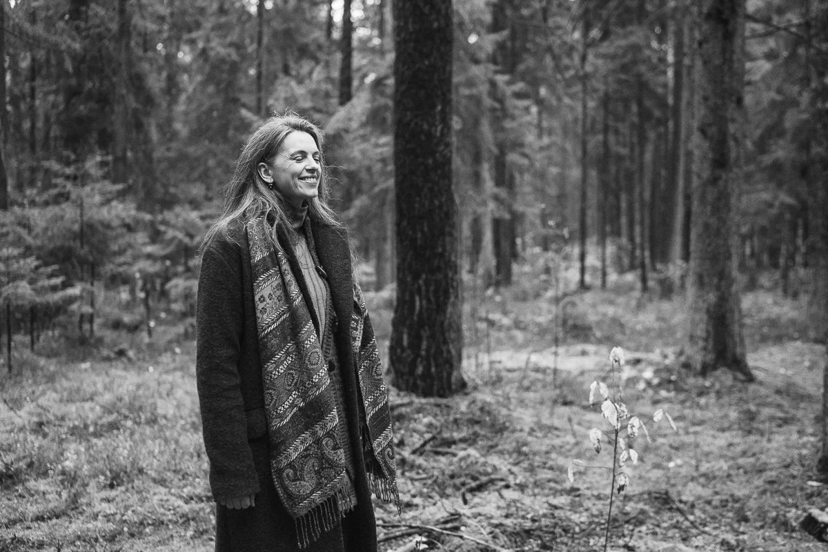 A woman smiling while walking in a forest with tall trees, wearing a dark jacket and a patterned scarf.