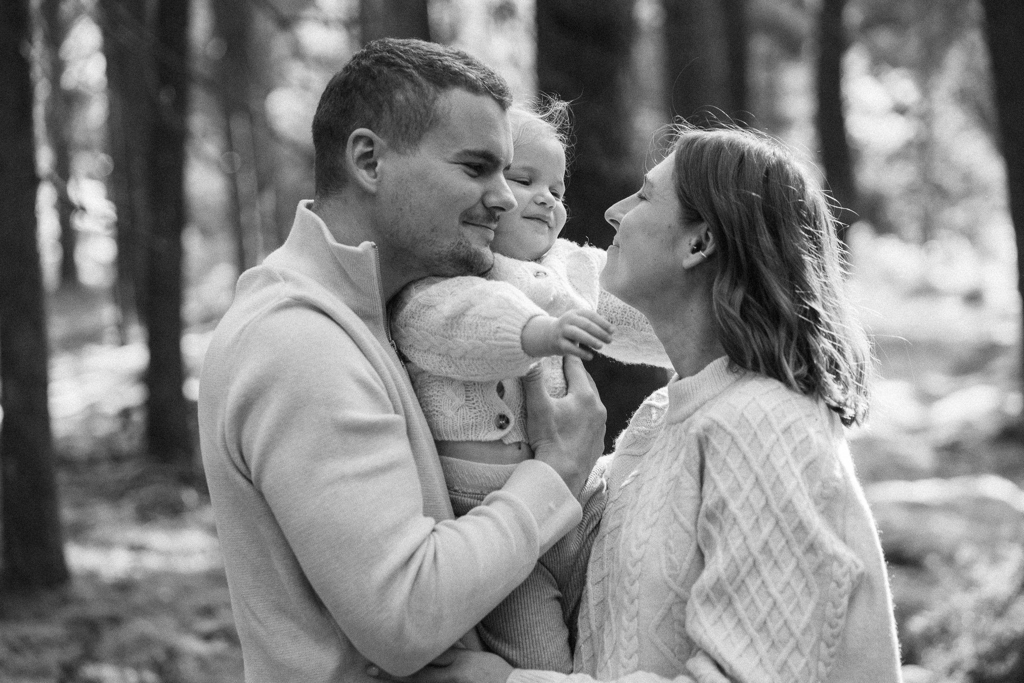 A black and white photo of a happy family outdoors in a wooded area. The father and mother are embracing their young daughter, who is smiling with her eyes closed as her parents lean in to kiss her on each side of her face.