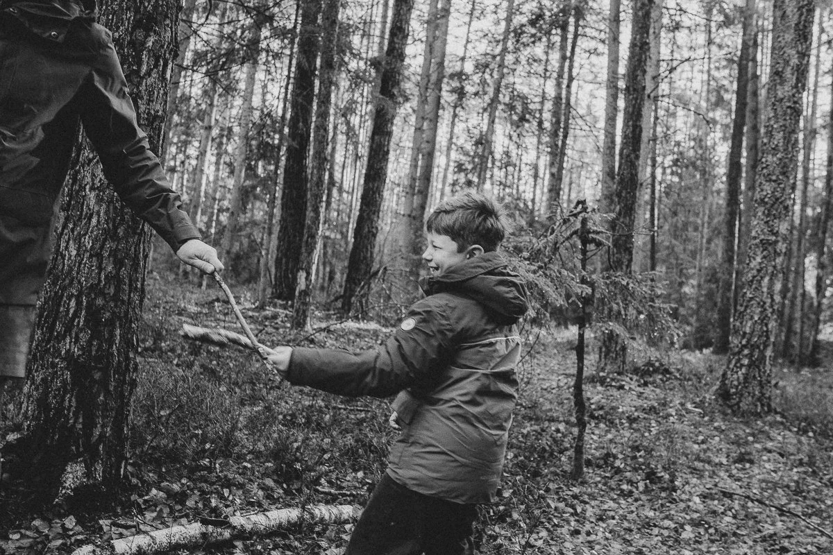 A young boy in a jacket pulling on a stick held by an adult in a forest.