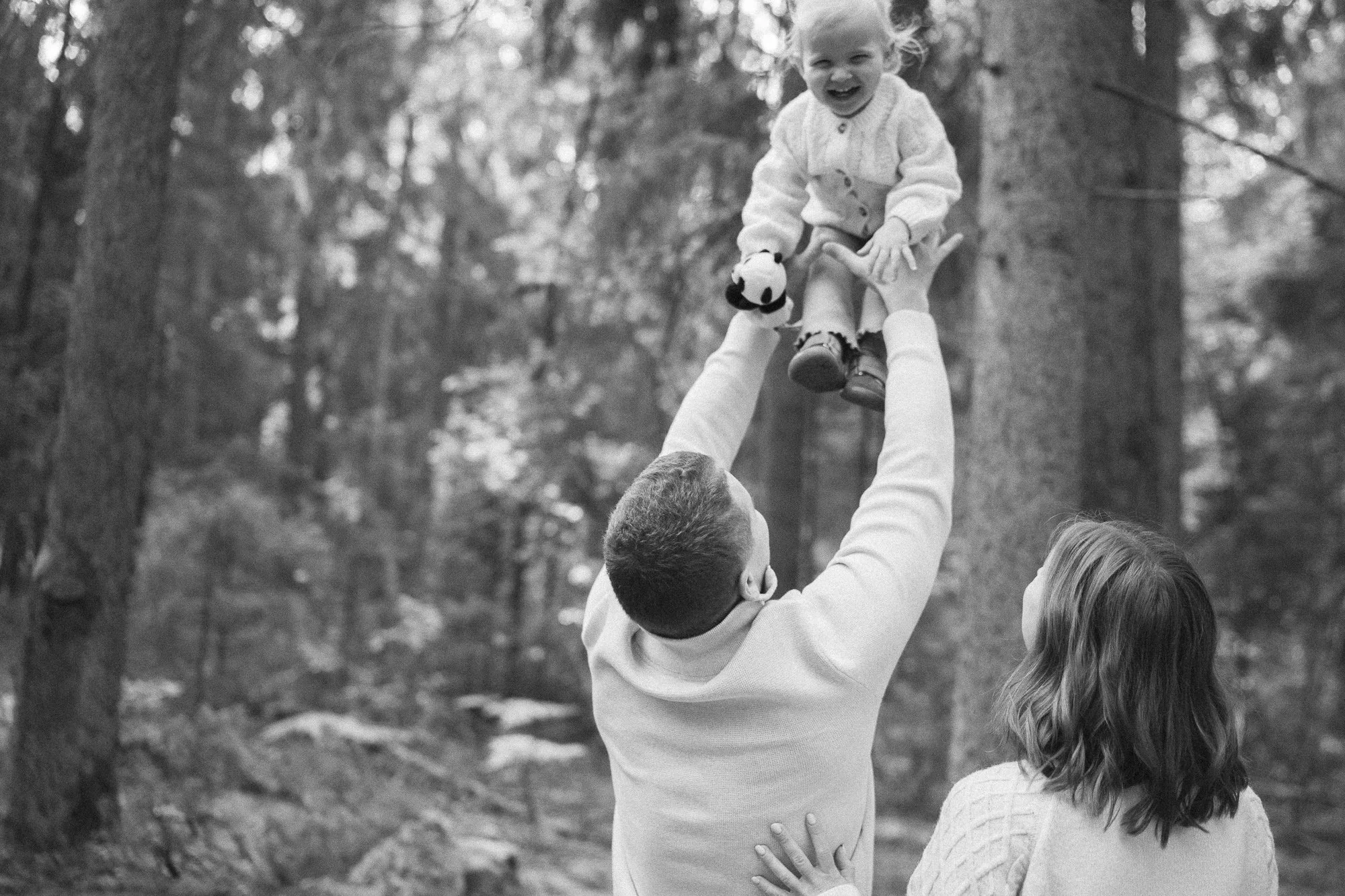 A man lifting a young girl in the air outdoors in a wooded area, with another girl standing nearby, both in white sweaters.