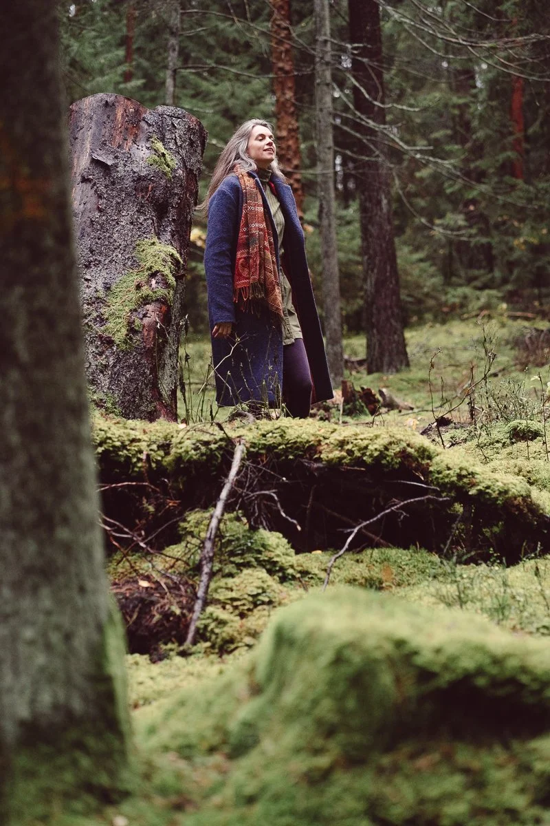 A woman with long gray hair, wearing a dark coat and a colorful scarf, stands in a dense forest surrounded by tall trees and moss-covered forest floor. She appears to be enjoying the peaceful natural environment.
