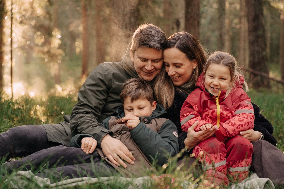 Family of four sitting together on a blanket in a forest during sunset, smiling and embracing each other.