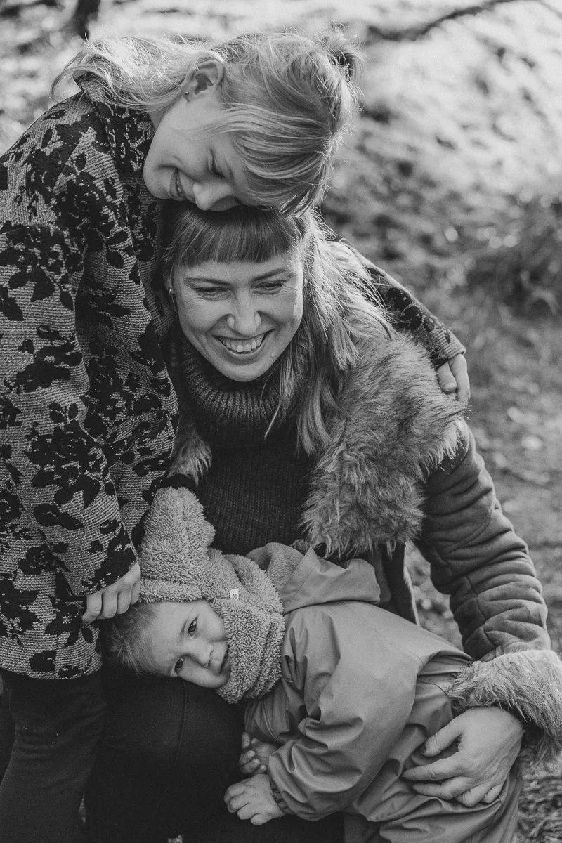 A woman and a young girl are hugging and smiling outdoors. The woman has curly hair and the girl is wearing a coat and a fuzzy hat. They appear happy and affectionate.