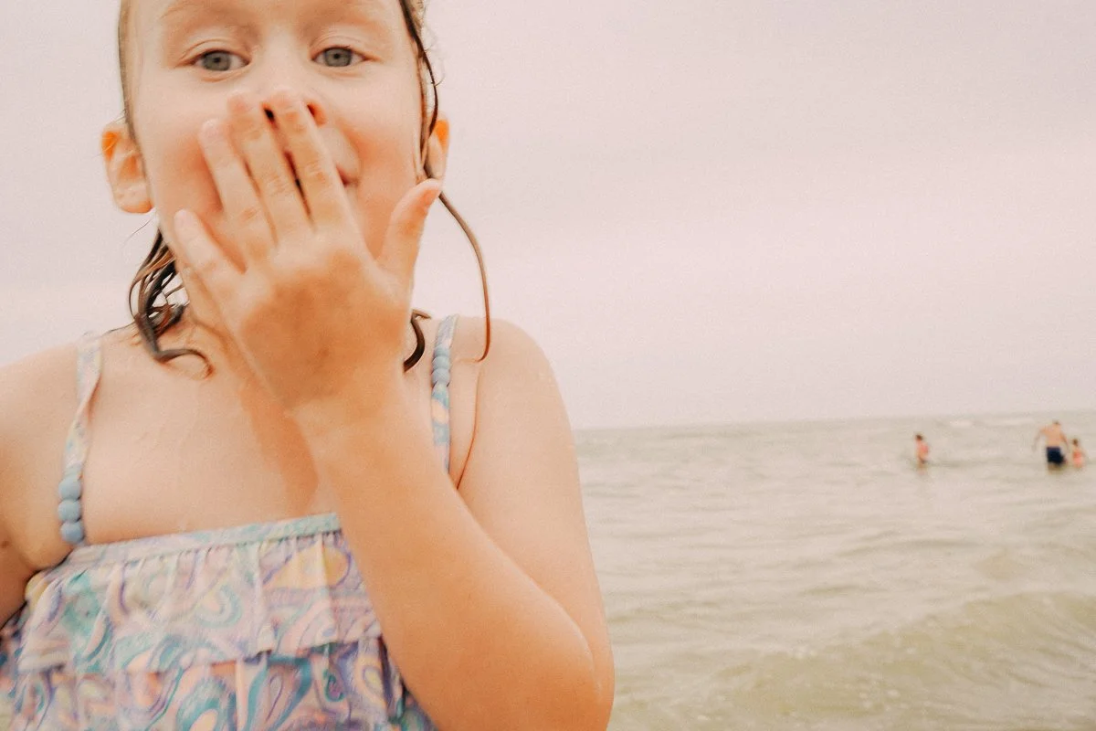 A young girl at the beach with wet hair, wearing a patterned swimsuit, blowing a kiss with her hand covering her mouth, while in the background there are people swimming in the ocean.
