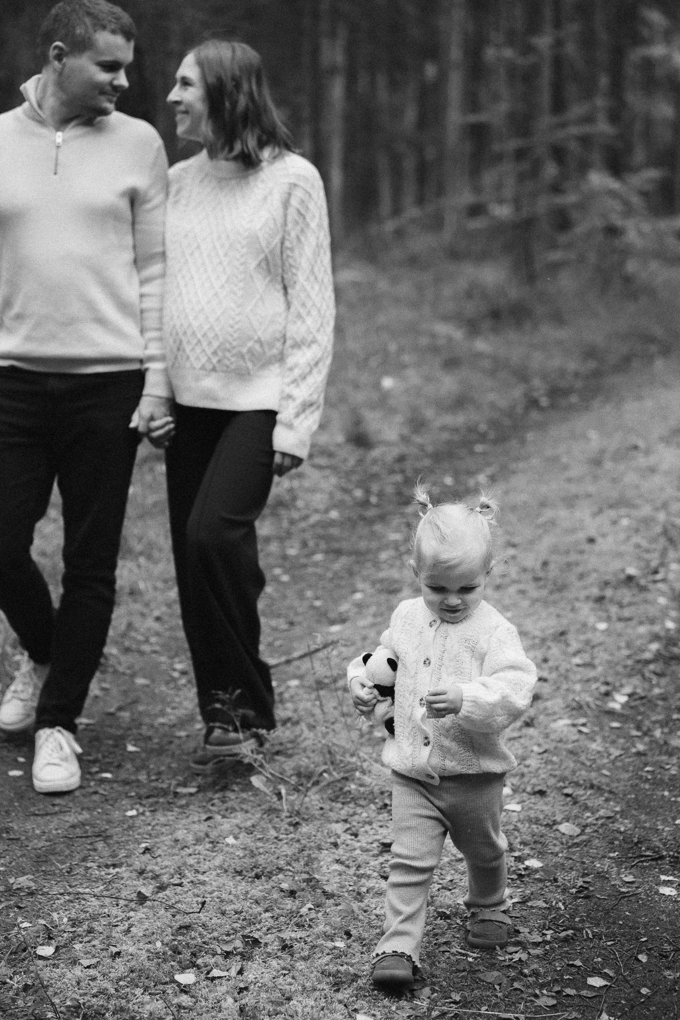 A black and white photo of a family walk in a forest. A man and woman are holding hands on the left side of the image, while a young girl with light hair tied in pigtails, wearing a sweater and pants, is walking in front of them, holding a stuffed an