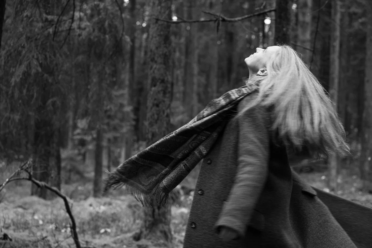 A woman with long hair throws back her head and looks up as she walks through a forest, wearing a coat and a patterned scarf.