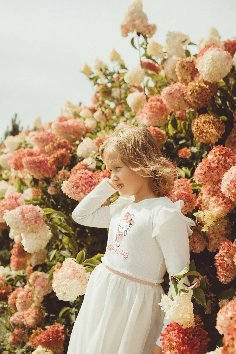 A young girl with curly blonde hair standing in front of a large bush of pink and white hydrangea flowers, smiling and looking to the side.