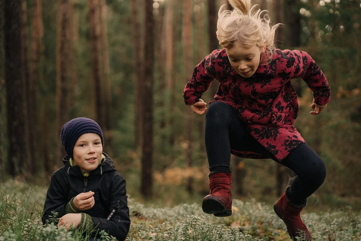 Two children playing outdoors in a forest, one girl jumping in the air and one boy sitting on the ground with a forest background.