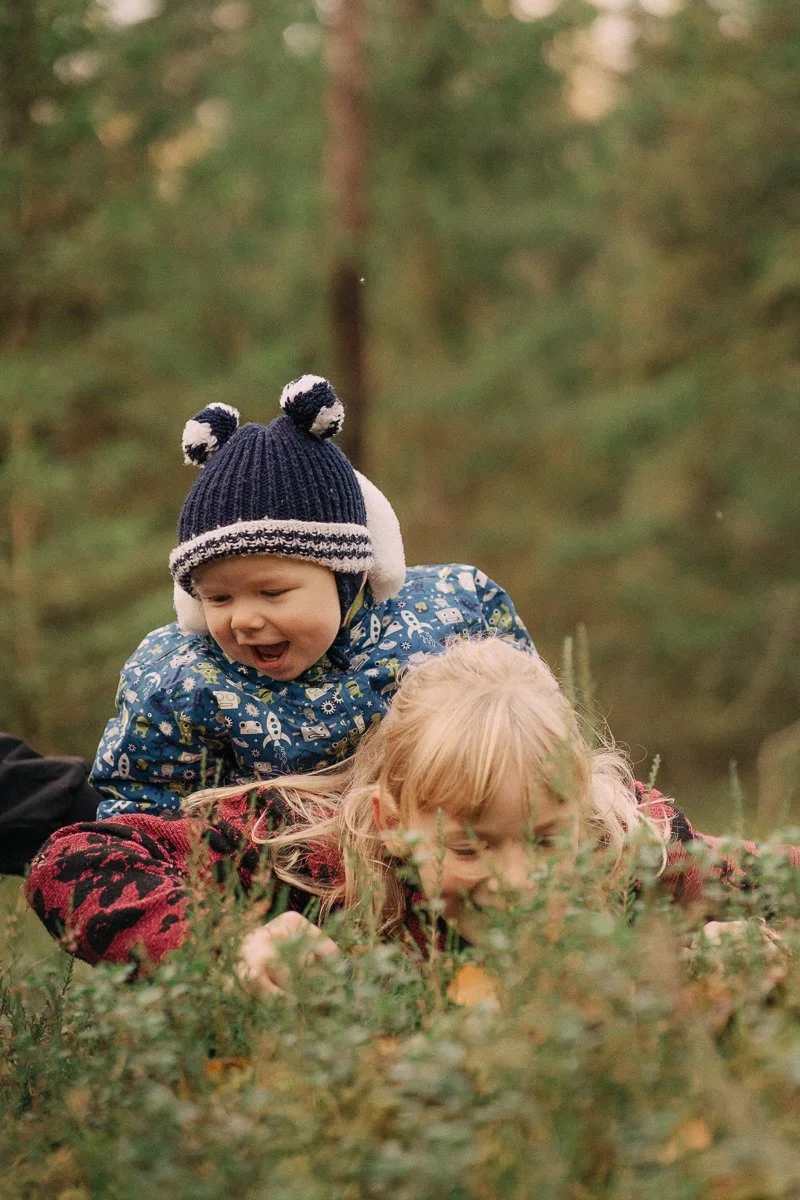 Two children playing outdoors in a forest. One boy with a blue winter hat with pom-poms smiling while climbing on top of a girl with blonde hair in a red and black sweater. The background shows green trees and blurred nature.