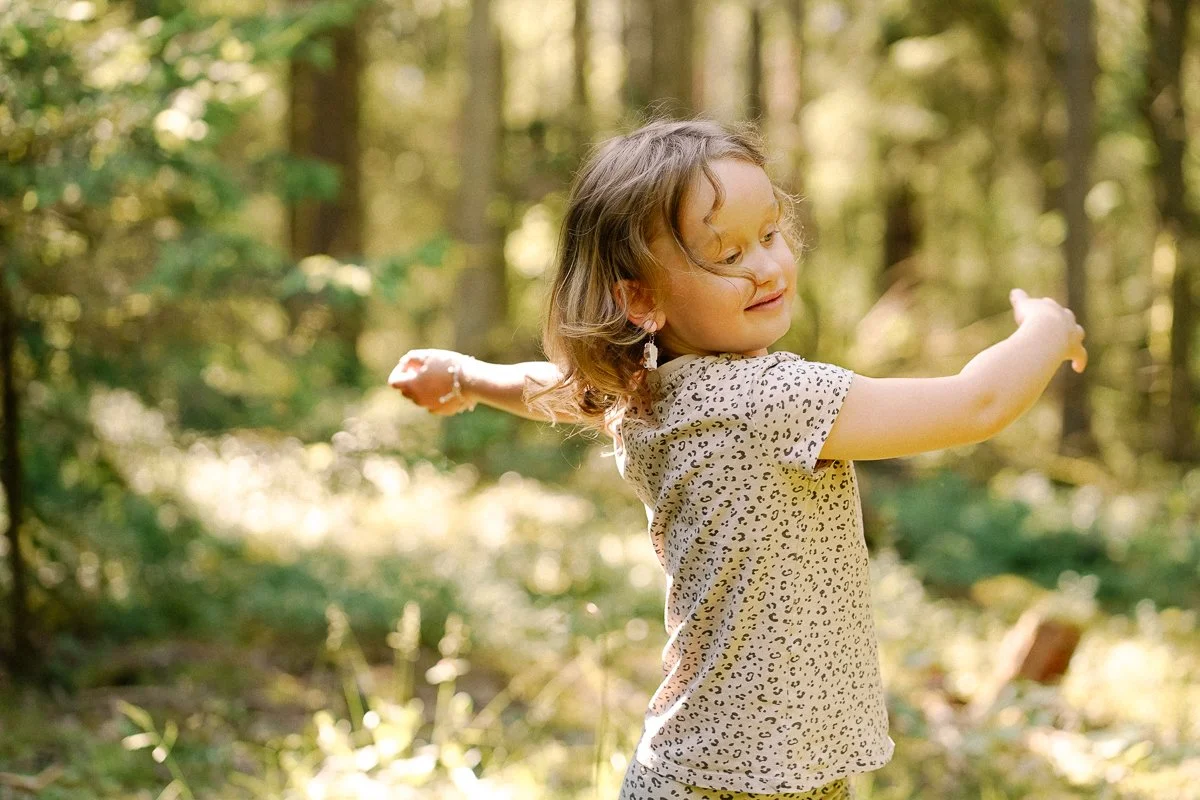 Young girl with curly hair and earrings in a forest, smiling and extending her arms.