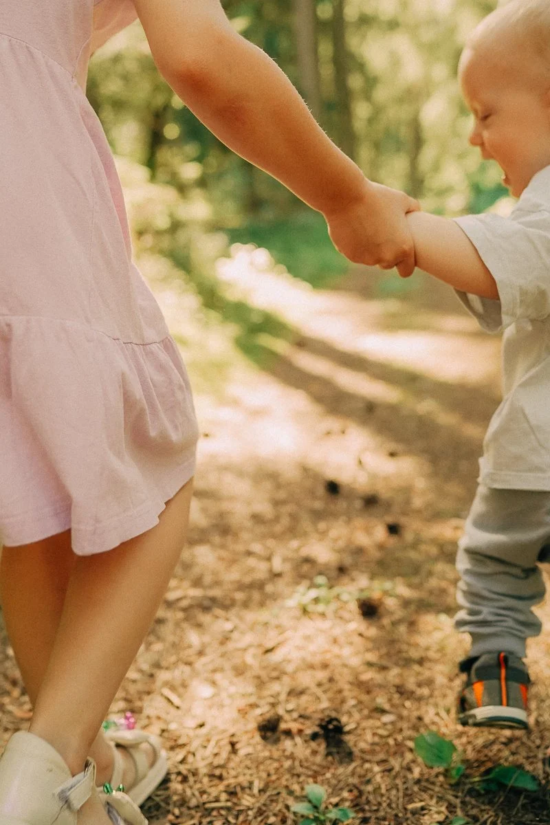 A woman and a young child holding hands while walking on a forest trail.