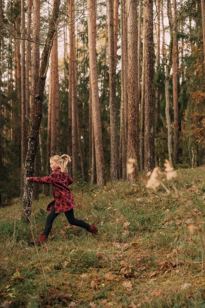 A young girl with a ponytail, wearing a red and black coat, black pants, and red shoes, is running through a forest with tall trees and fallen leaves.