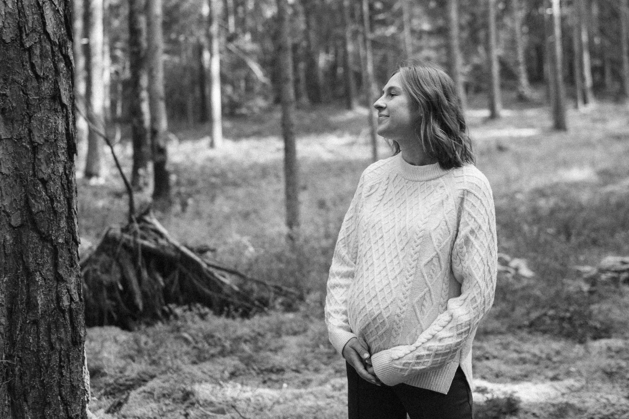 A woman with shoulder-length hair wearing a knitted sweater standing outdoors in a forest, looking at a tree with a calm expression.