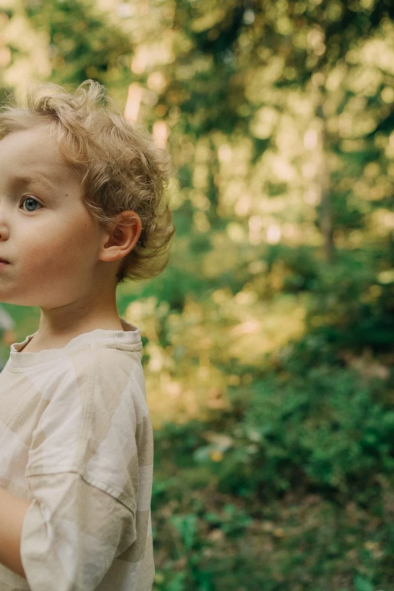 A young boy with curly blonde hair and blue eyes standing outdoors in a wooded area.