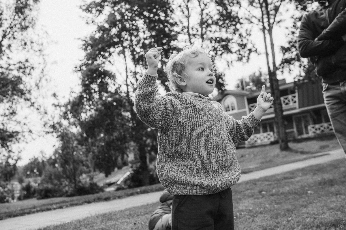 A young boy with curly hair, wearing a sweater, standing outdoors with trees and a building in the background, raising his hands and looking up with an expressive face.