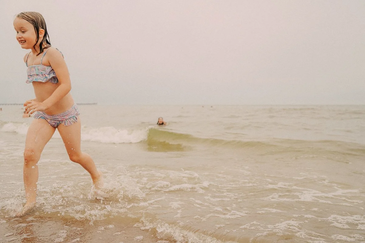 A young girl running and playing in the ocean at the beach, with a person swimming in the background.