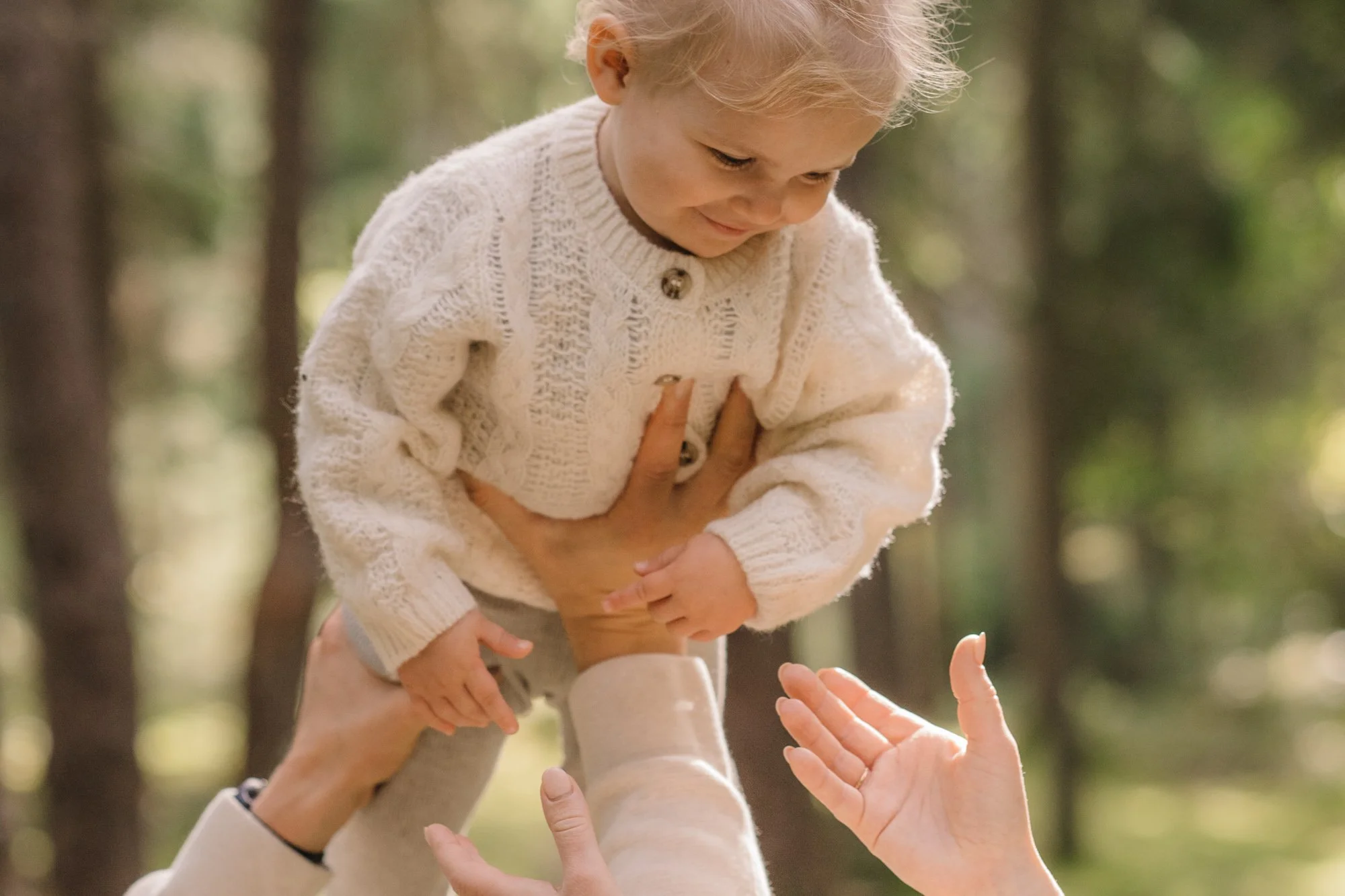 A young girl with blonde hair smiling while being lifted in the air outdoors, holding her hand over her chest and reaching out to hands below.