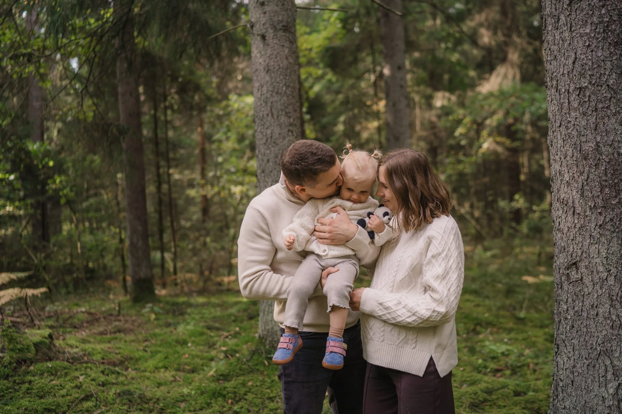 A family of three in a forest, with a man holding a young girl and the woman touching her, all smiling and interacting closely.