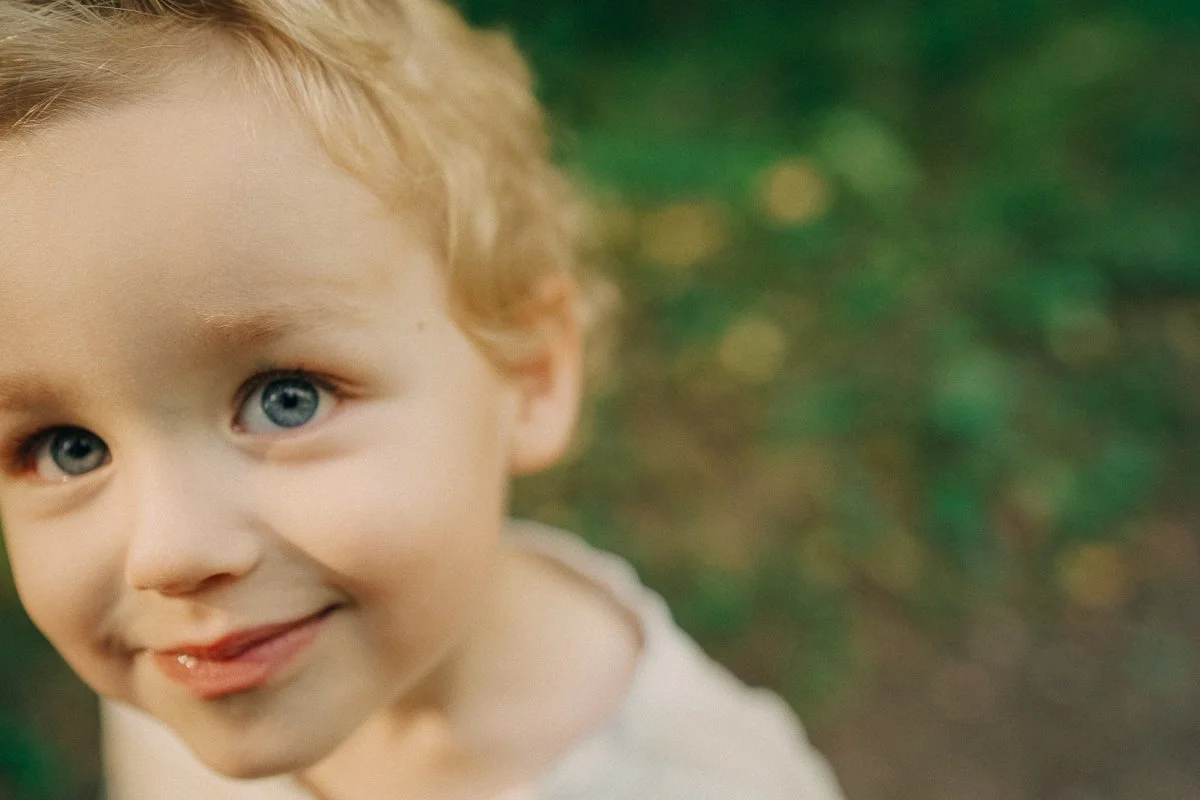 Close-up of a smiling young boy with blue eyes and blonde curly hair, outdoors with blurred green background.