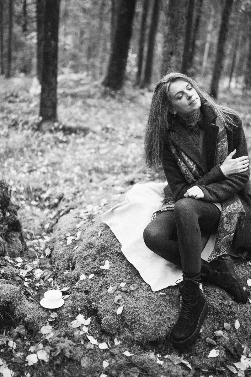 A woman with long hair sitting on a rock in a forest with her eyes closed, surrounded by trees and fallen leaves, in black and white.