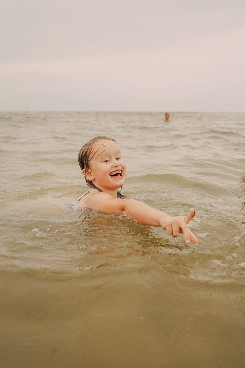 A young girl swimming and smiling in the ocean with waves around her, pointing forward with her finger.