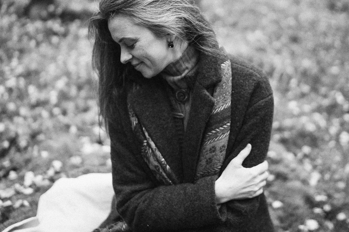 A woman with long hair, wearing a coat and patterned scarf, sitting outdoors on the ground, with arms crossed and looking downward.